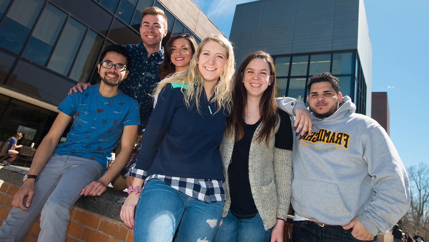 Diverse group of smiling students leaning against a brick wall outside.